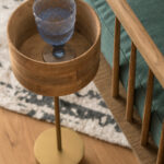 Detail eines Beistelltischs aus Holz und Messing mit blauem Stielglas, neben grünem Sessel und Teppich