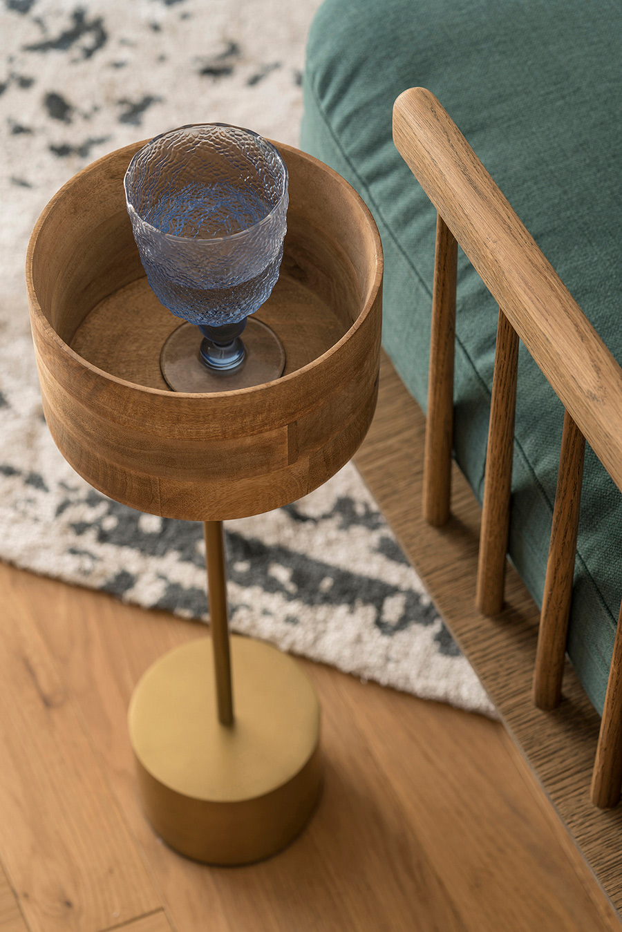 Detail eines Beistelltischs aus Holz und Messing mit blauem Stielglas, neben grünem Sessel und Teppich