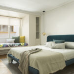 Schlafzimmer mit petrolblauem Polsterkopfteil, Sitzbank am Fenster mit gemusterten Kissen und Einbauschränken