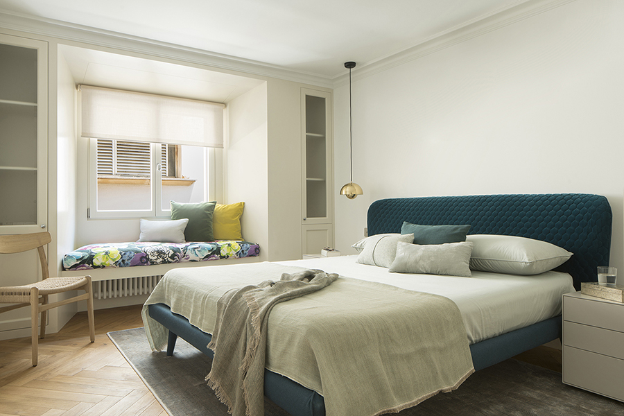 Schlafzimmer mit petrolblauem Polsterkopfteil, Sitzbank am Fenster mit gemusterten Kissen und Einbauschränken