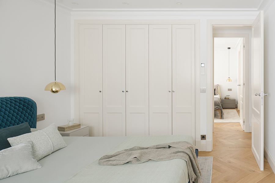 Dormitorio con armarios blancos empotrados de puertas clásicas, cabecero azul petróleo y lámpara dorada colgante