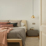 Schlafzimmer mit Holzbett und beigem Kopfteil, rosa und grauen Decken, grauem Nachttisch und Messingleuchte
