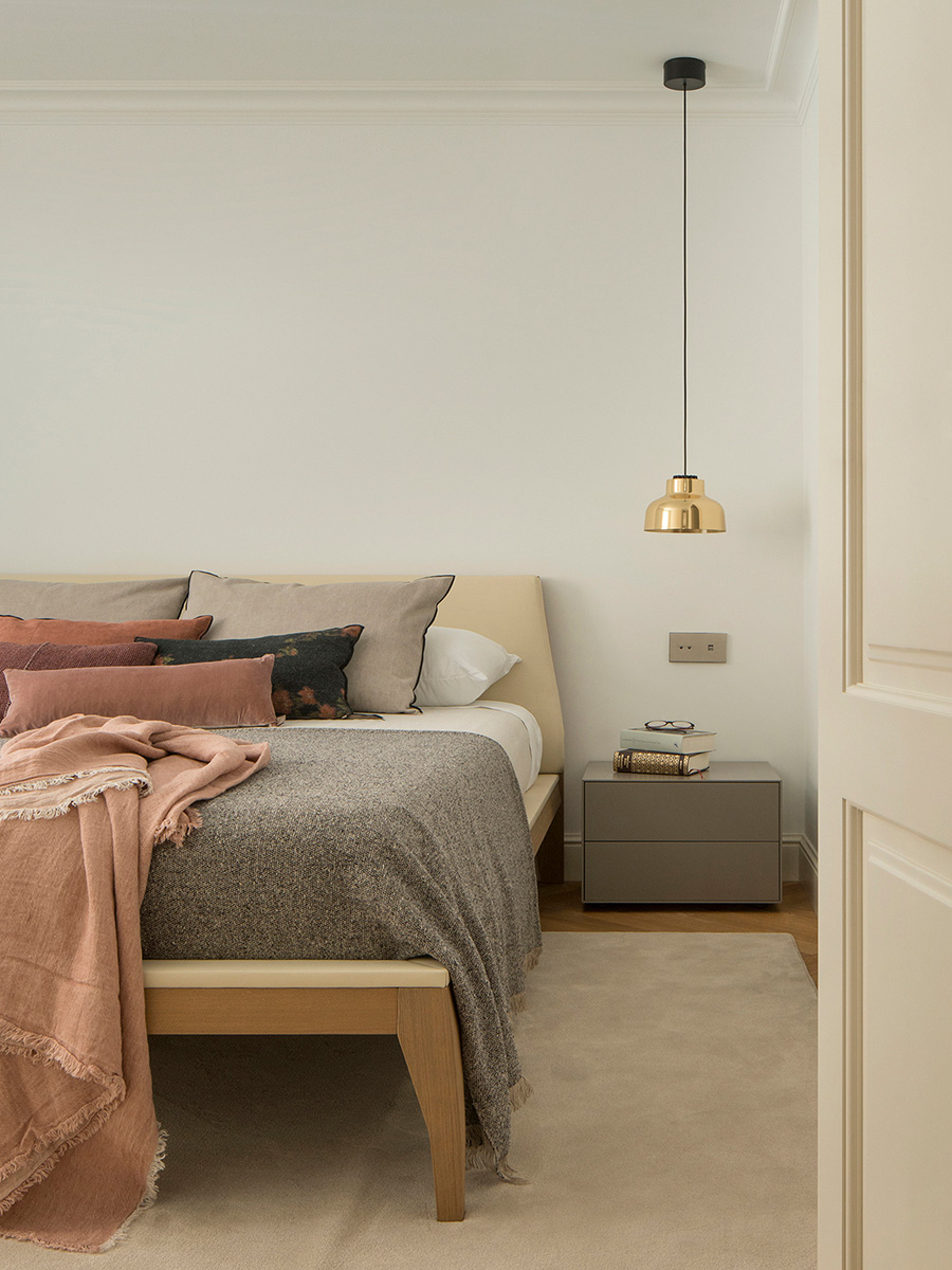 Schlafzimmer mit Holzbett und beigem Kopfteil, rosa und grauen Decken, grauem Nachttisch und Messingleuchte