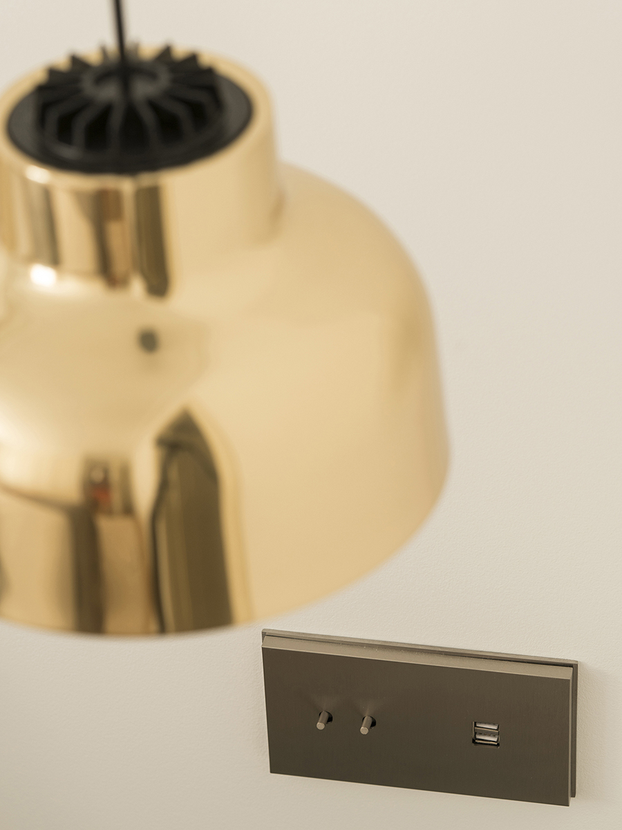 Detail einer Pendelleuchte aus poliertem Messing und metallener Schalterblende mit Schaltern und USB-Steckdose an der Wand