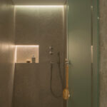 Mit grauem Stein verkleidete Dusche mit beleuchteter Nische, Metallarmaturen, Deckenbrause und transluzenter Duschwand