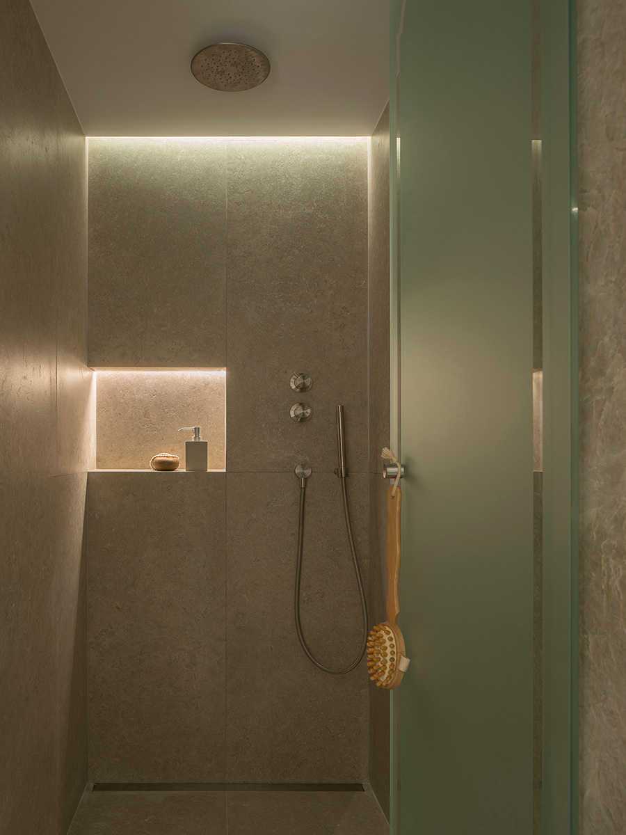 Mit grauem Stein verkleidete Dusche mit beleuchteter Nische, Metallarmaturen, Deckenbrause und transluzenter Duschwand