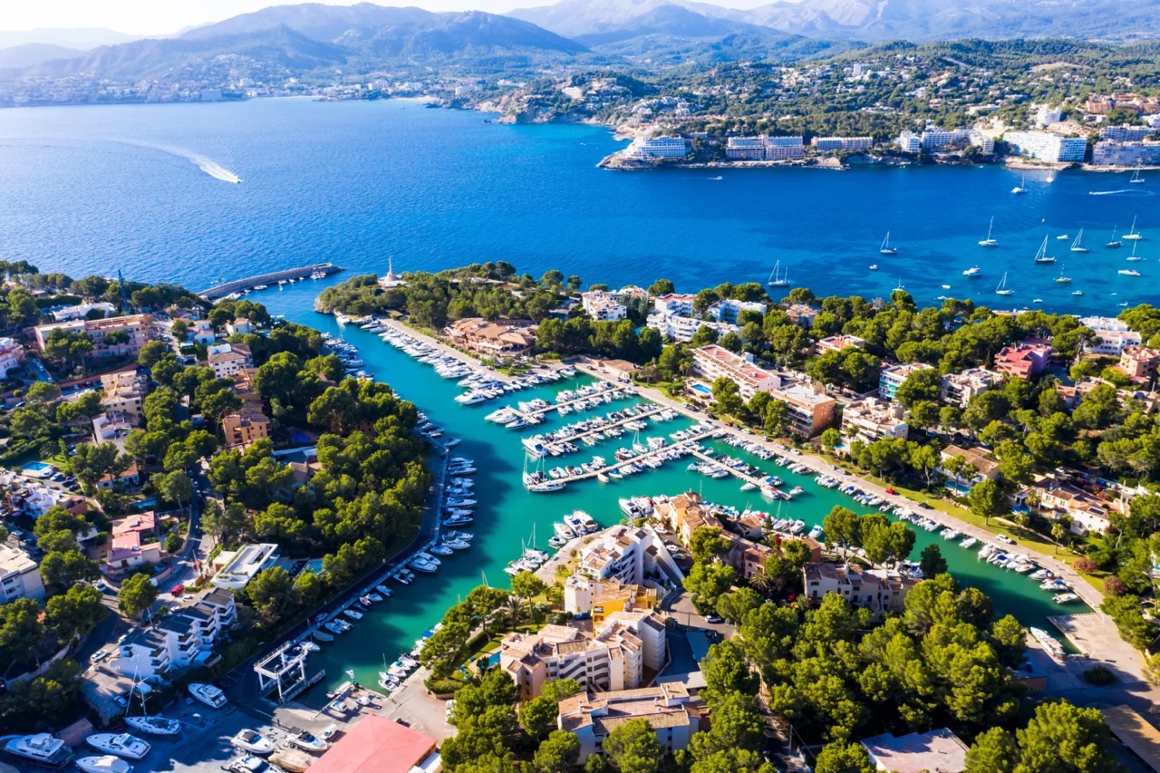 Luftaufnahme eines Yachthafens mit Segelbooten, umgeben von Pinien und Wohnsiedlungen an der Bucht