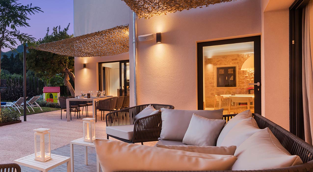 Zona de estar exterior con sofá trenzado y cojines bajo toldo, comedor al aire libre y jardín