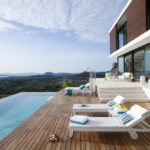 Blick vom Holzdeck entlang des Pools, mit Sonnenliegen und Bergen am Horizont