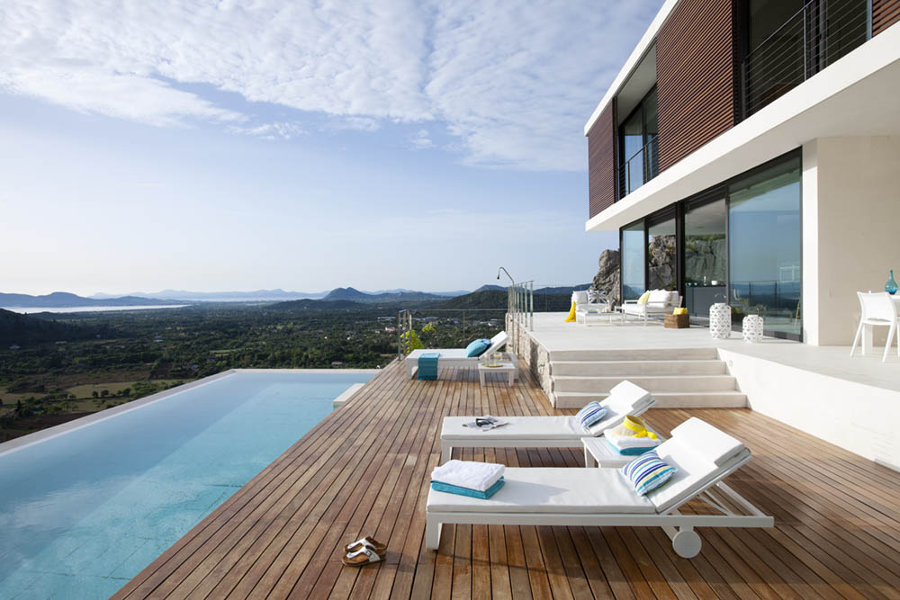 Blick vom Holzdeck entlang des Pools, mit Sonnenliegen und Bergen am Horizont