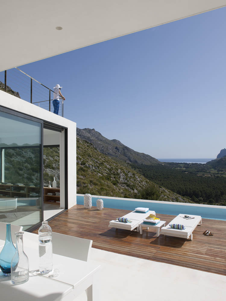 Terrasse mit Holzdeck, Sonnenliegen und Pool, eine Person lehnt am Geländer mit Blick ins Tal