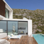 Weiße Fassade mit großen Fenstern, Holzdeck, Sonnenliegen und Pool vor dem felsigen Hang