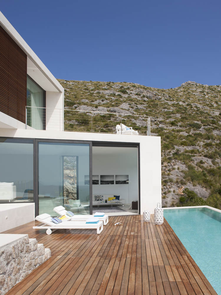 Weiße Fassade mit großen Fenstern, Holzdeck, Sonnenliegen und Pool vor dem felsigen Hang