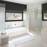 Badezimmer mit eingebauter Badewanne, Dusche aus Milchglas, Fenster mit dunklem Rollo und weißem Badvorleger