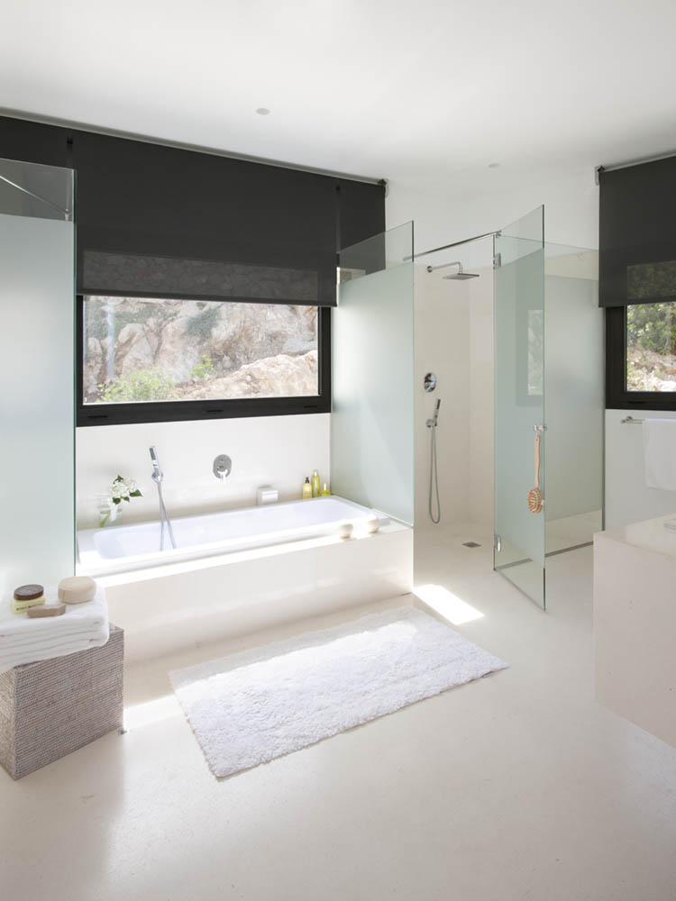 Badezimmer mit eingebauter Badewanne, Dusche aus Milchglas, Fenster mit dunklem Rollo und weißem Badvorleger