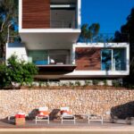 Vista frontal de la casa desde la piscina, con tumbonas, muro de piedra y lamas de madera