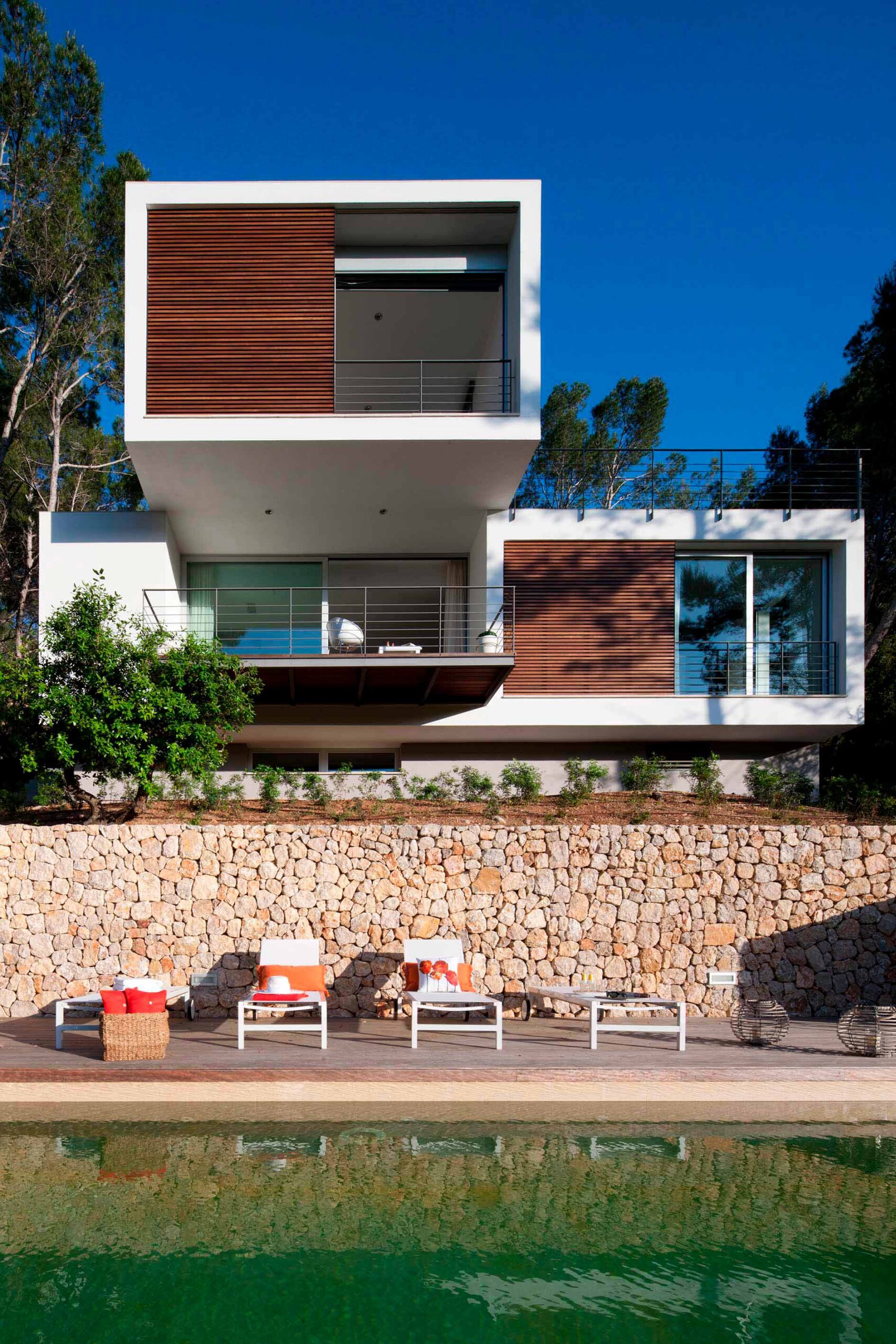 Vista frontal de la casa desde la piscina, con tumbonas, muro de piedra y lamas de madera