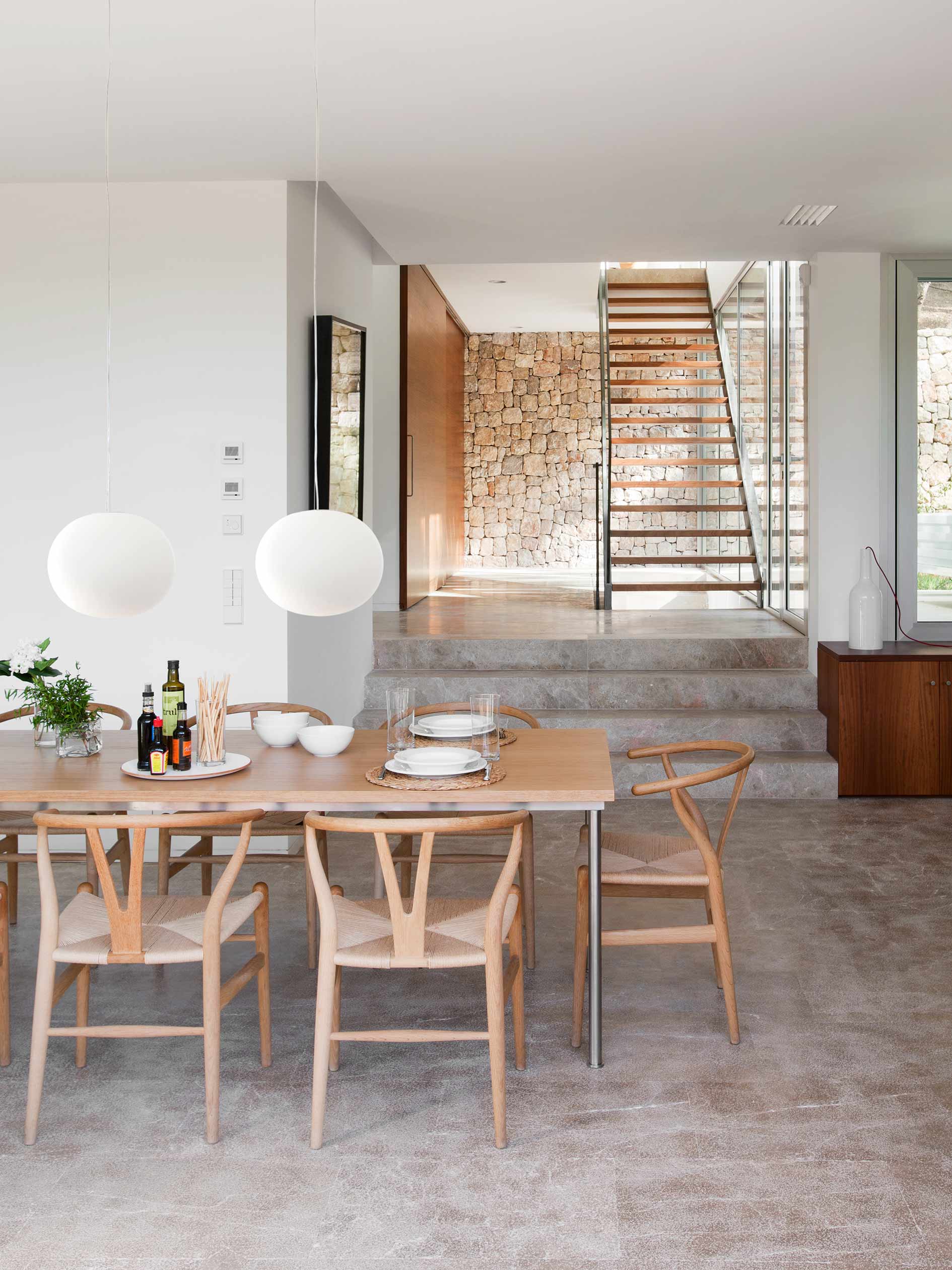 Detalle vertical del comedor: lámparas esféricas colgantes, sillas de madera y escalera con pared de piedra al fondo