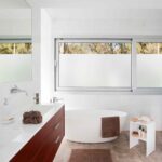 Baño luminoso con bañera exenta blanca, mueble de madera oscura, suelo de piedra y ventana al bosque