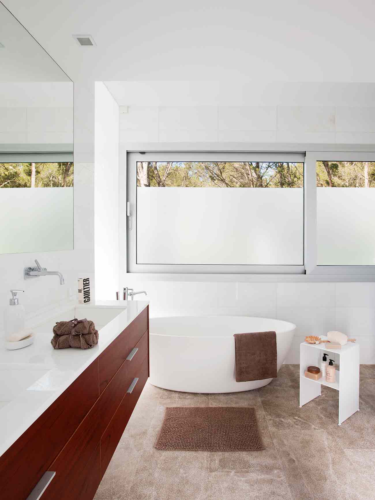 Baño luminoso con bañera exenta blanca, mueble de madera oscura, suelo de piedra y ventana al bosque