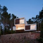 Fachada iluminada al anochecer sobre muros de piedra, con módulos blancos y lamas de madera entre pinos