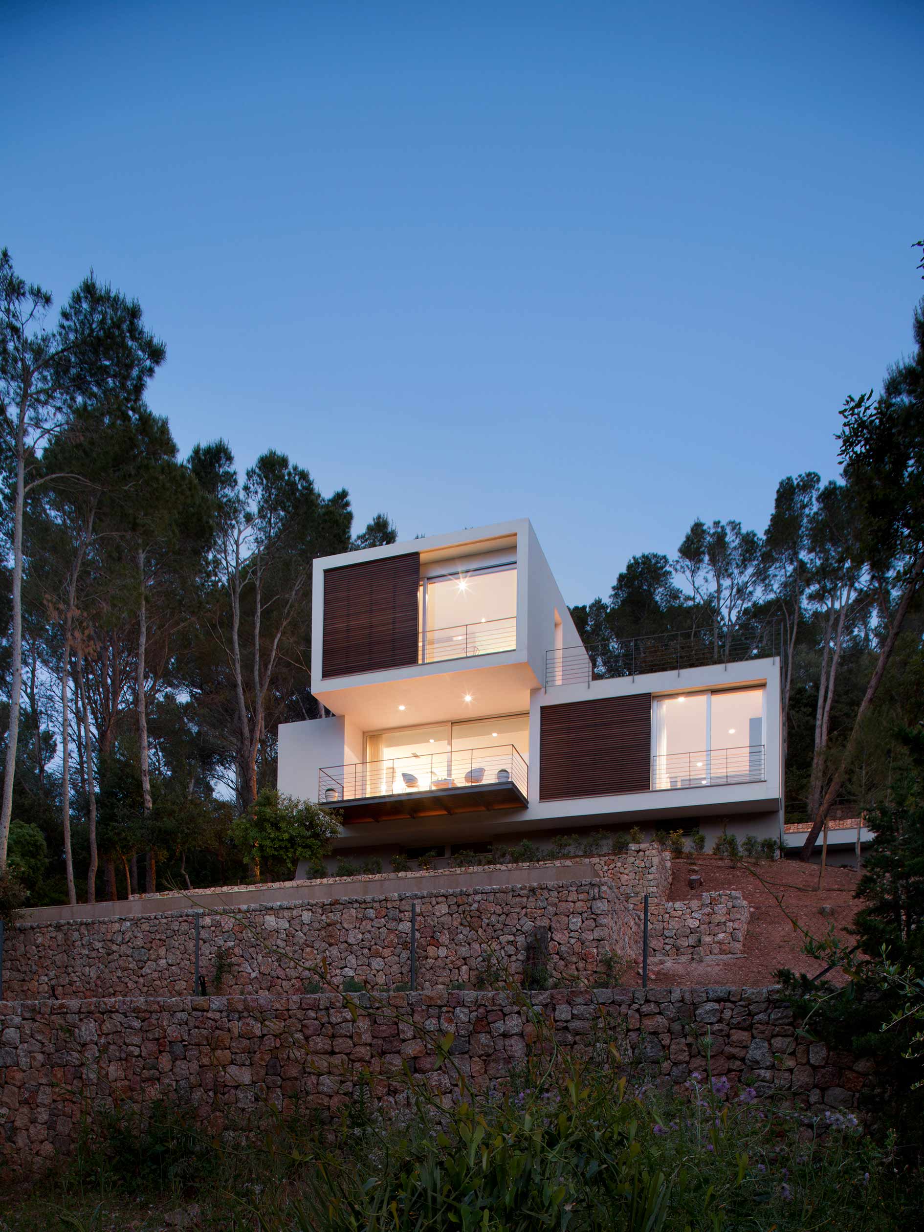 Fachada iluminada al anochecer sobre muros de piedra, con módulos blancos y lamas de madera entre pinos