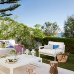 Terrasse unter blauem Himmel mit Kiefer, weißem Sessel, Weidenkorb, Hecke und dem Meer im Hintergrund
