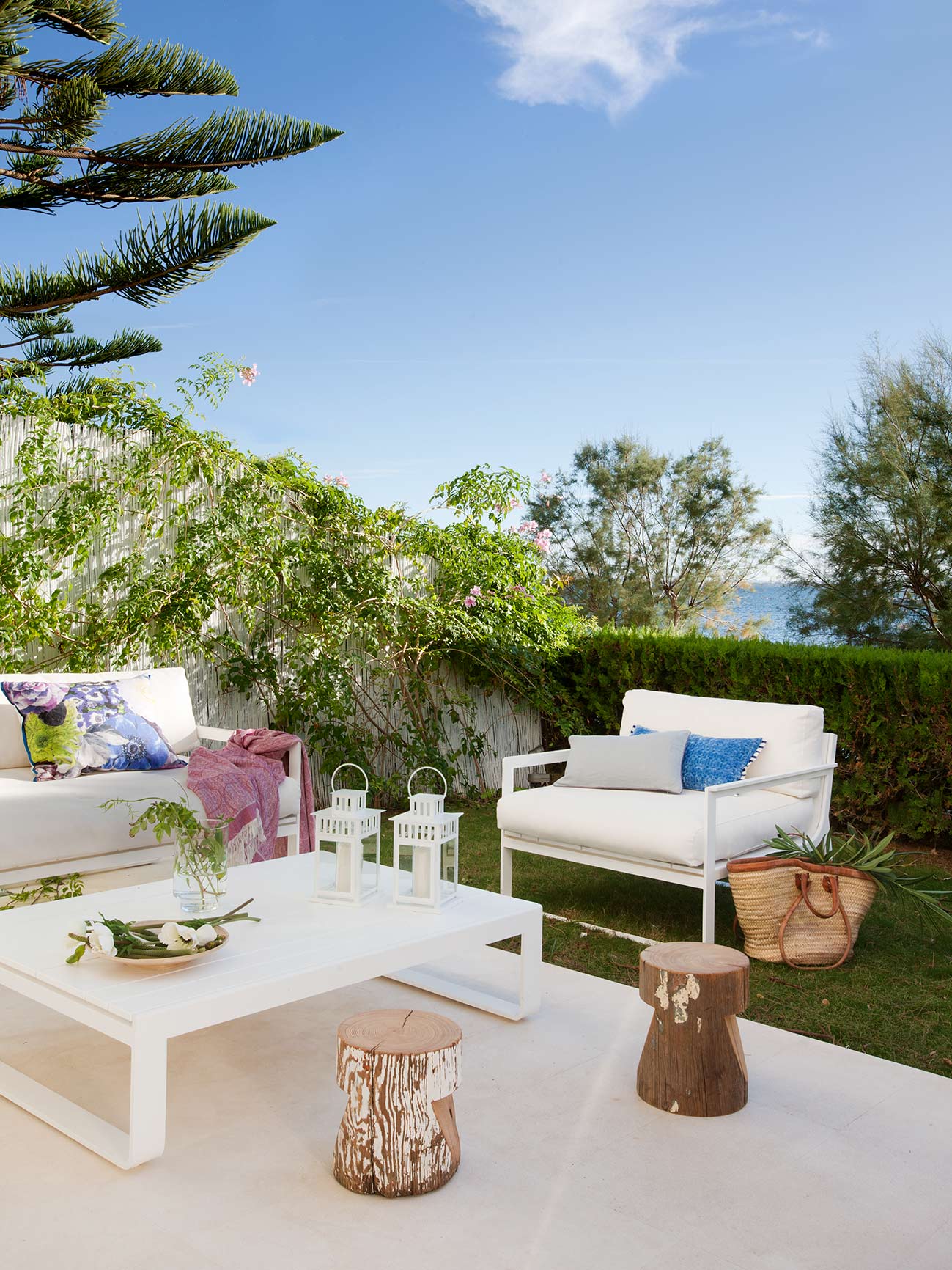 Terrasse unter blauem Himmel mit Kiefer, weißem Sessel, Weidenkorb, Hecke und dem Meer im Hintergrund