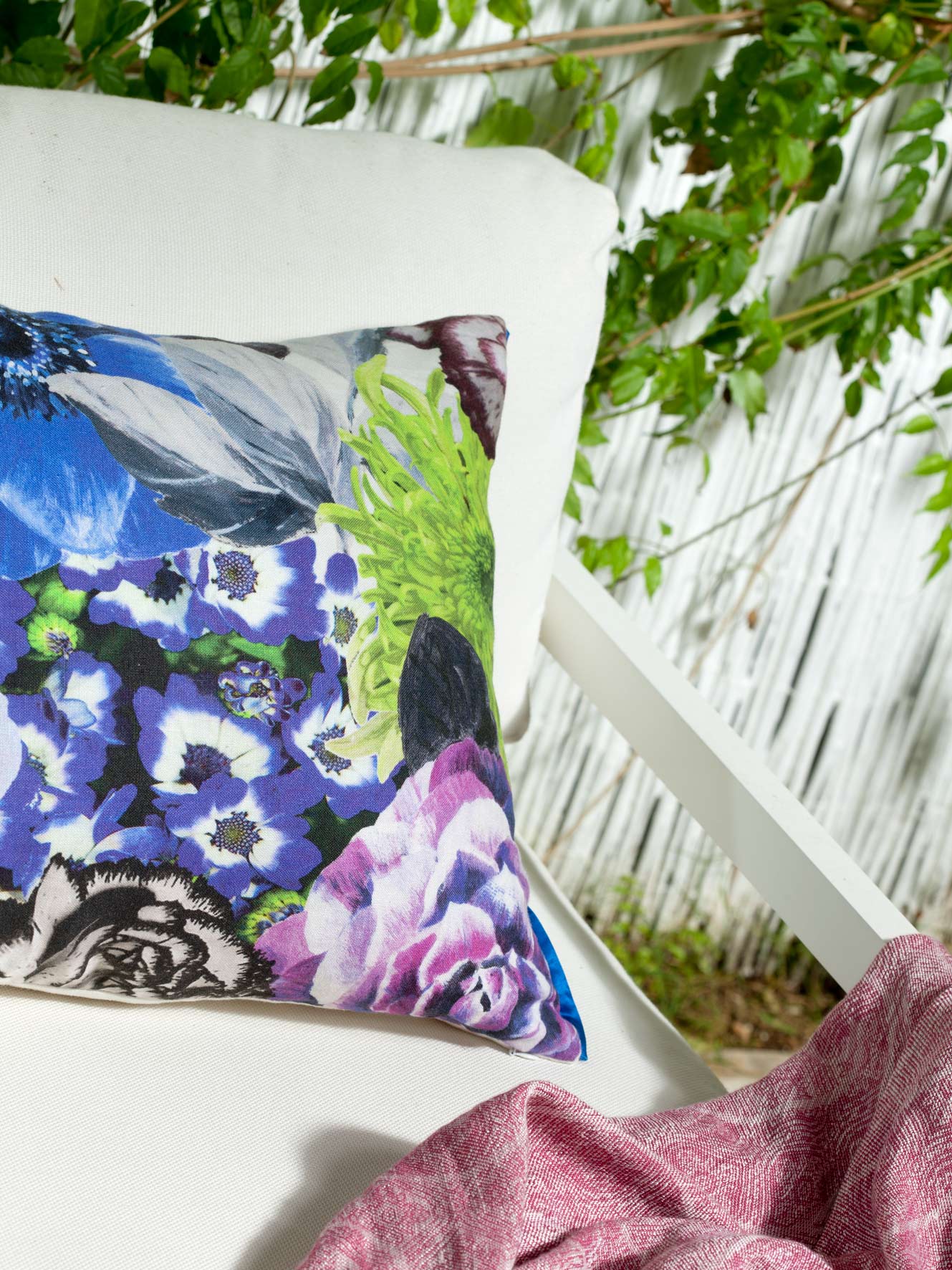 Geblümtes Kissen in Blau, Violett und Grün auf weißem Sessel, mit rosa Decke und Schilfzaun