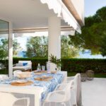 Mit Markise überdachte Terrasse, Tisch mit blau gemustertem Tischtuch und weißen Stühlen, dahinter Hecke und Meer