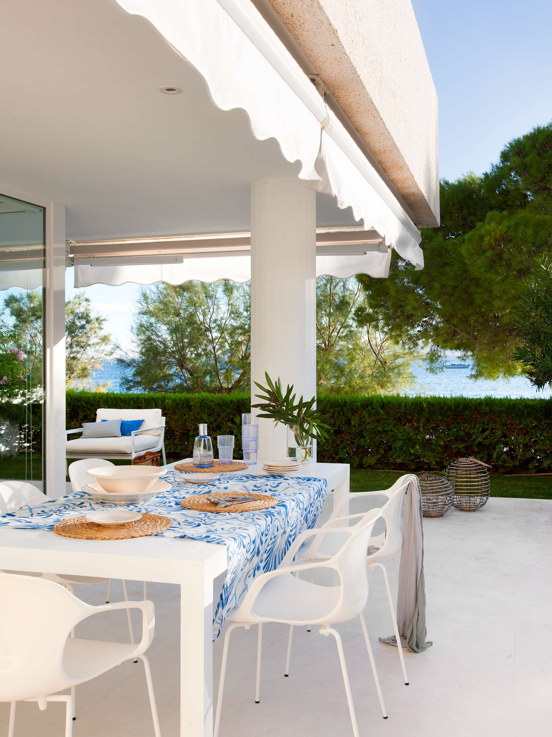 Mit Markise überdachte Terrasse, Tisch mit blau gemustertem Tischtuch und weißen Stühlen, dahinter Hecke und Meer