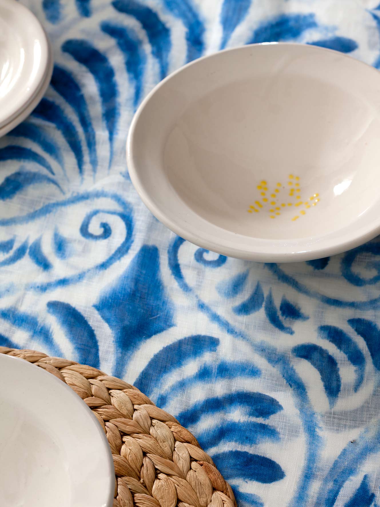 Detail eines blau-weißen Tischtuchs mit heller Keramikschale und Untersetzer aus geflochtener Faser