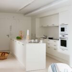 Weiße Küche mit Insel, glatten Schrankfronten, eingebautem Backofen und Mikrowelle, hellem Boden und Weidenkorb