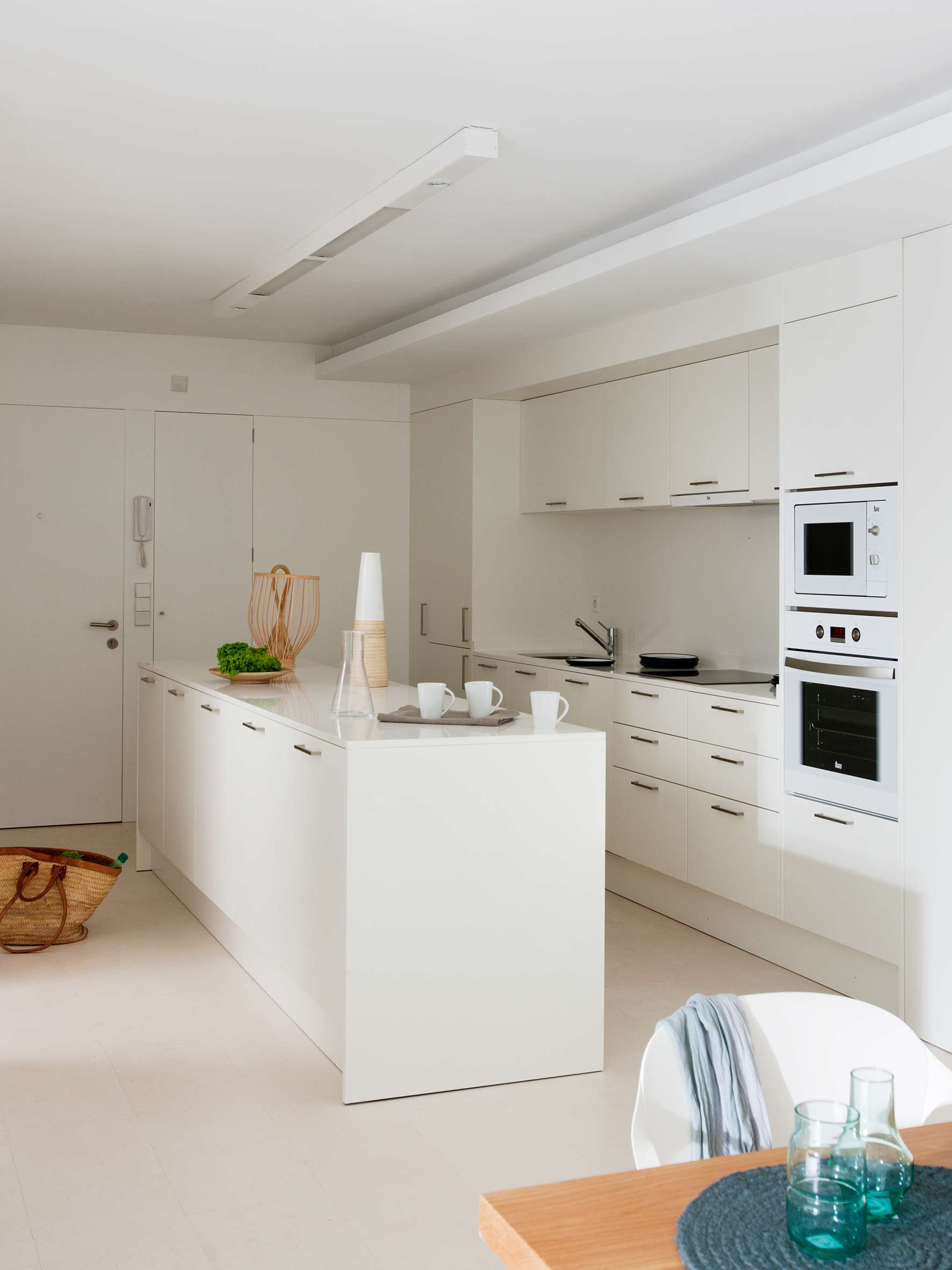 Weiße Küche mit Insel, glatten Schrankfronten, eingebautem Backofen und Mikrowelle, hellem Boden und Weidenkorb