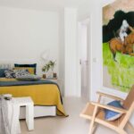 Weißes Schlafzimmer mit senfgelber Tagesdecke, Nachttisch, großem Reiterbild und Sessel aus geflochtenem Holz