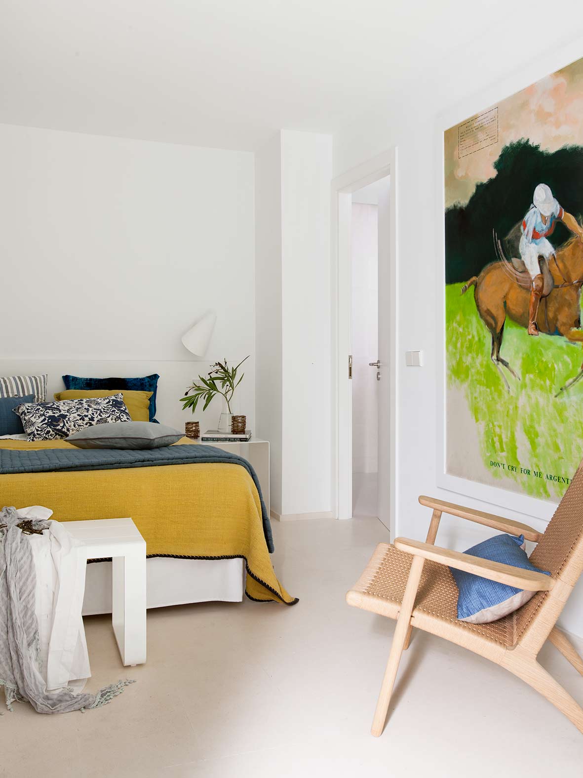 Weißes Schlafzimmer mit senfgelber Tagesdecke, Nachttisch, großem Reiterbild und Sessel aus geflochtenem Holz