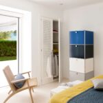 Schlafzimmer mit weißem Lamellenschrank, blau-grauem Modulmöbel, Sessel und großem Fenster zum Garten