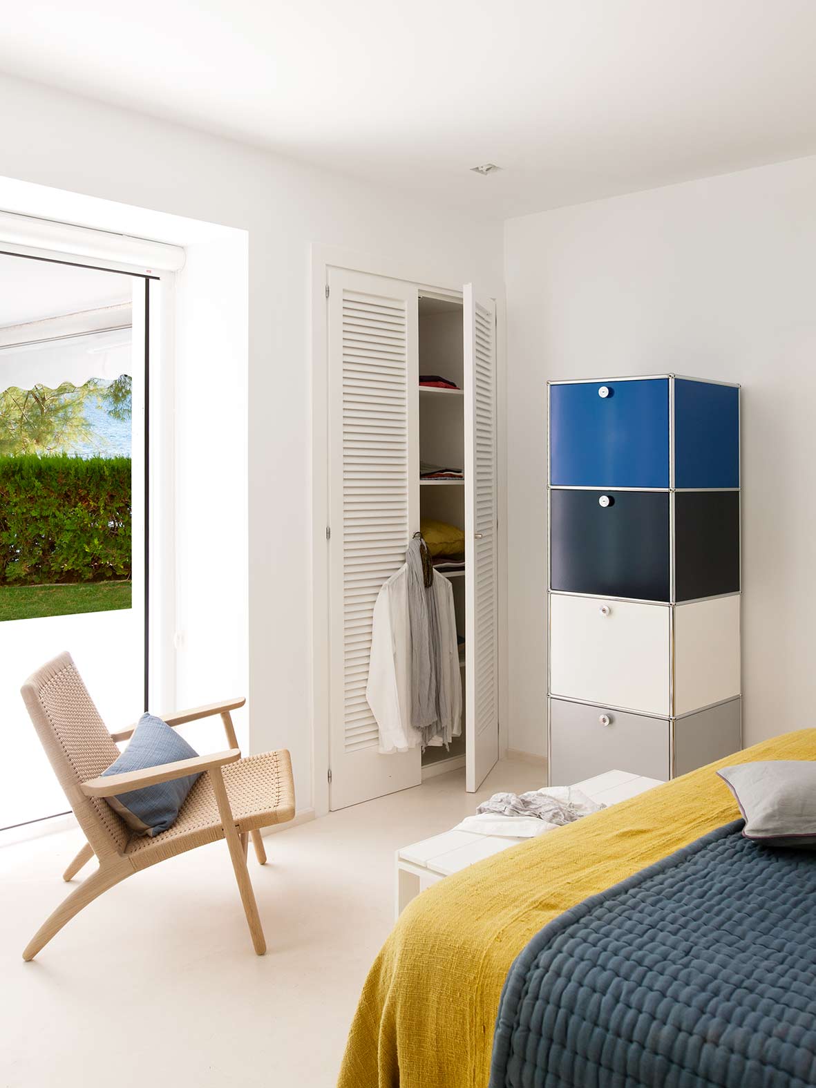 Schlafzimmer mit weißem Lamellenschrank, blau-grauem Modulmöbel, Sessel und großem Fenster zum Garten