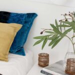 Detail eines blauen Samtkissens und eines senfgelben Kissens neben weißem Nachttisch mit Büchern und grünen Zweigen