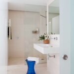 Badezimmer in Sandtönen mit Dusche hinter Glaswand, wandhängendem weißem Waschbecken und blauem Hocker