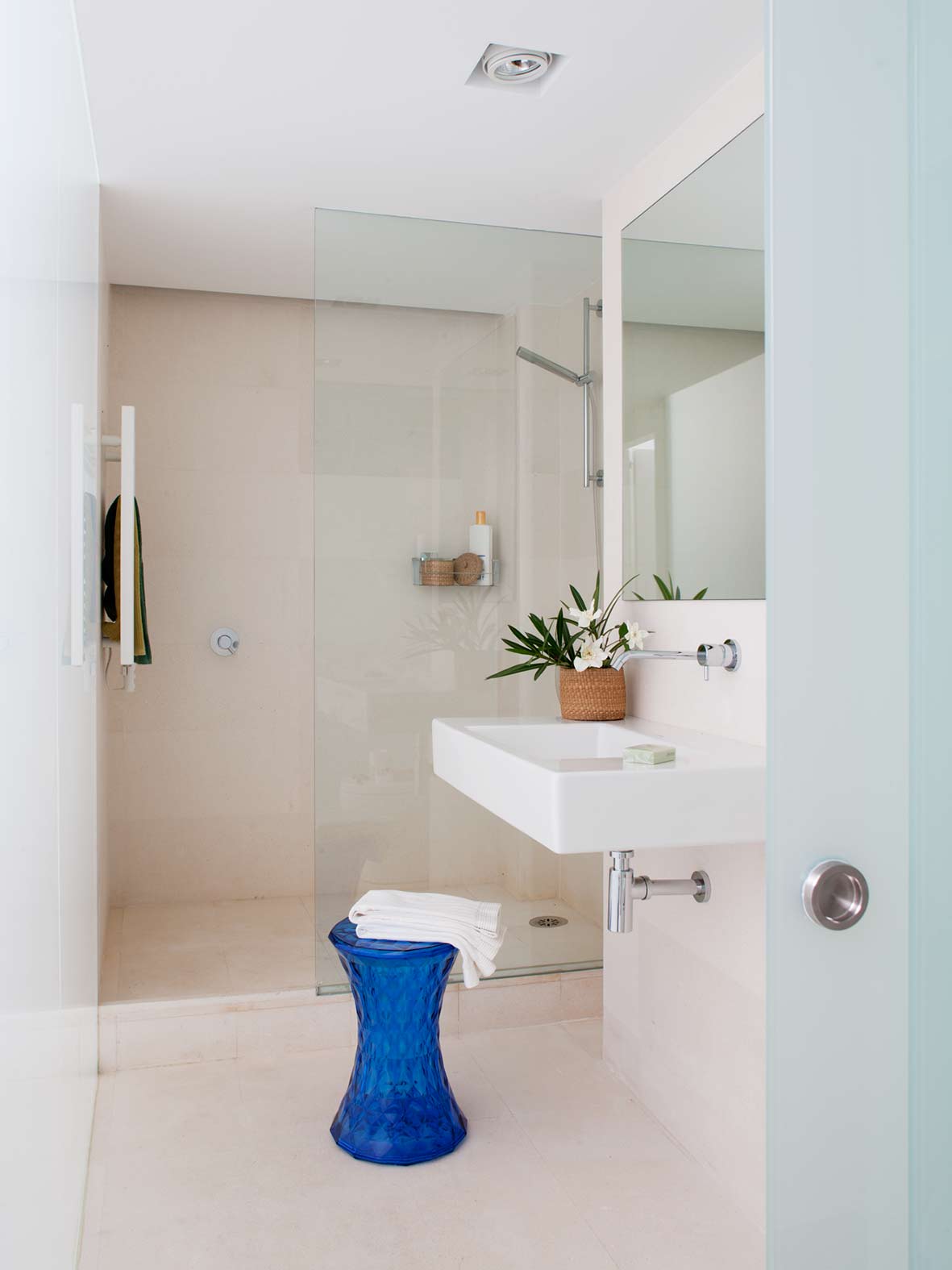 Badezimmer in Sandtönen mit Dusche hinter Glaswand, wandhängendem weißem Waschbecken und blauem Hocker