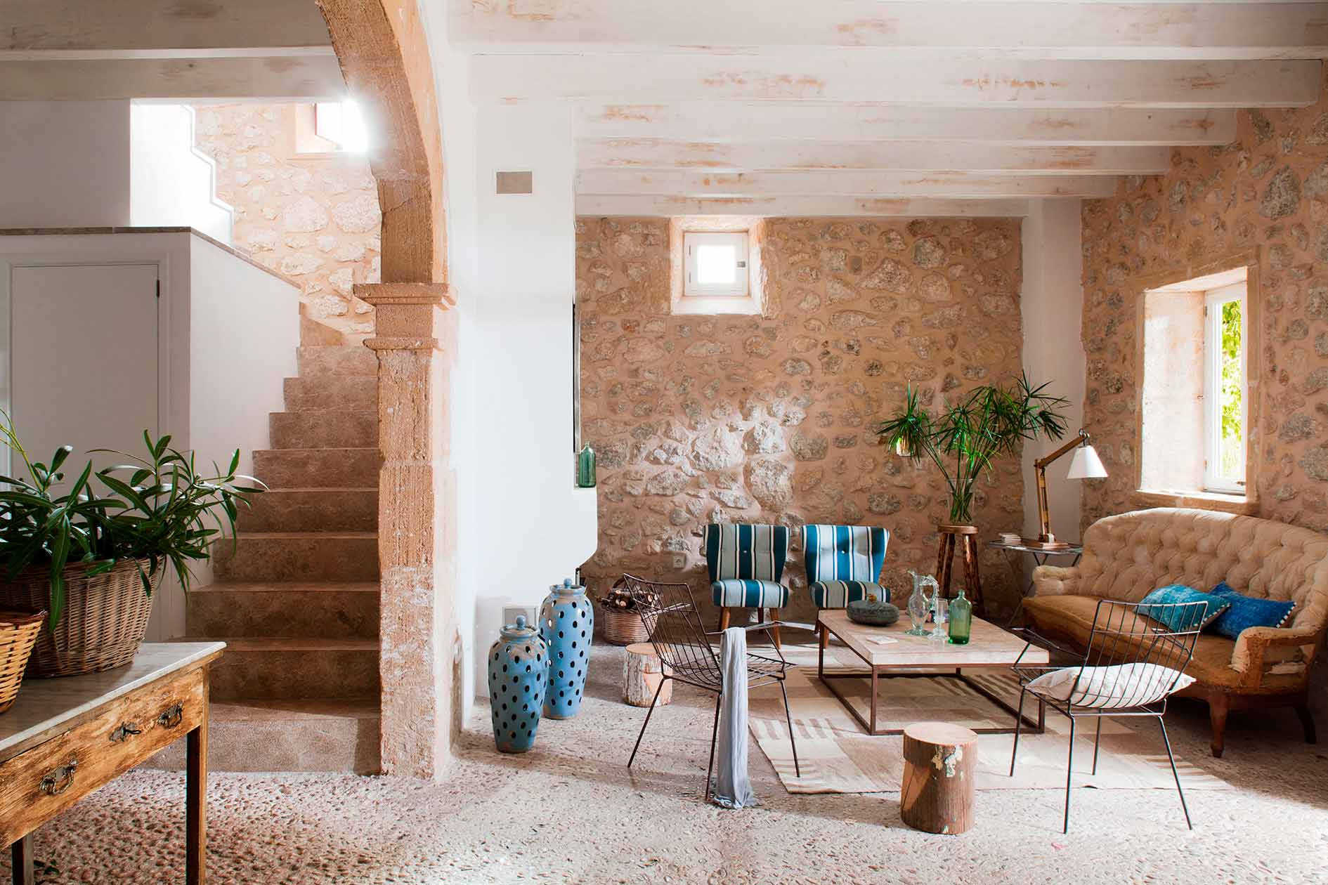 Salón con suelo de cantos rodados, arco de piedra, escalera lateral, vigas blancas y muros de mampostería