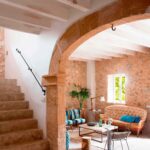 Escalera de piedra junto a un arco que enmarca el salón, con vigas blancas y sofá capitoné