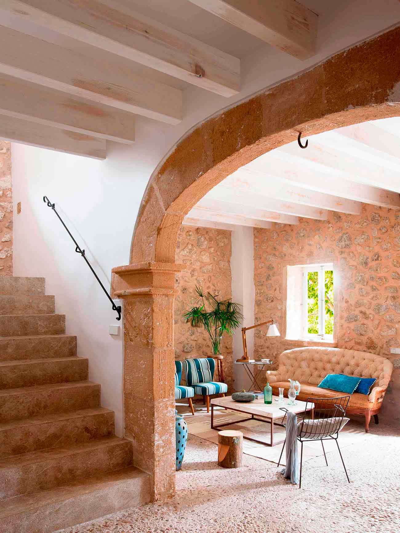 Escalera de piedra junto a un arco que enmarca el salón, con vigas blancas y sofá capitoné
