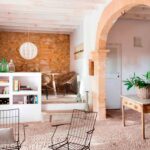 Sala con estantería blanca, comedor elevado al fondo, sillas de alambre y suelo empedrado