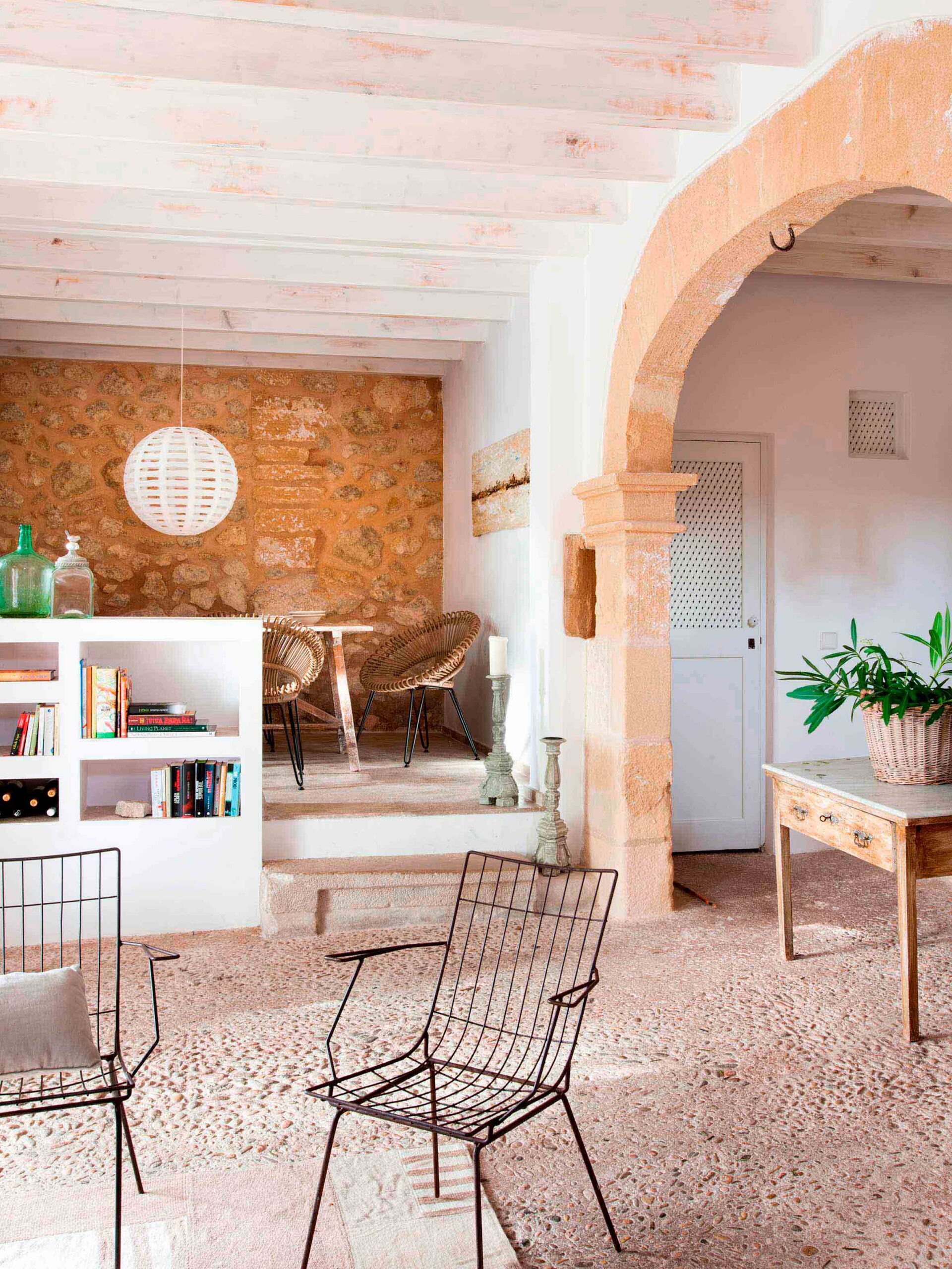 Sala con estantería blanca, comedor elevado al fondo, sillas de alambre y suelo empedrado
