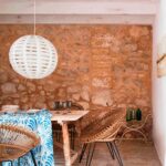 Comedor rústico con mesa de madera envejecida, sillas de mimbre, lámpara esférica blanca y muro de piedra