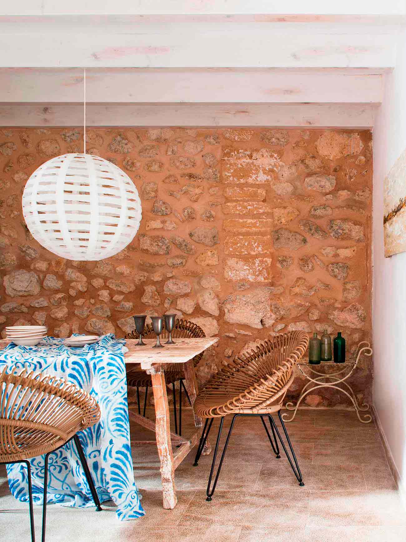 Comedor rústico con mesa de madera envejecida, sillas de mimbre, lámpara esférica blanca y muro de piedra