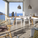 Vista desde el salón hacia el comedor con cristalera panorámica, mesa de madera y mar azul