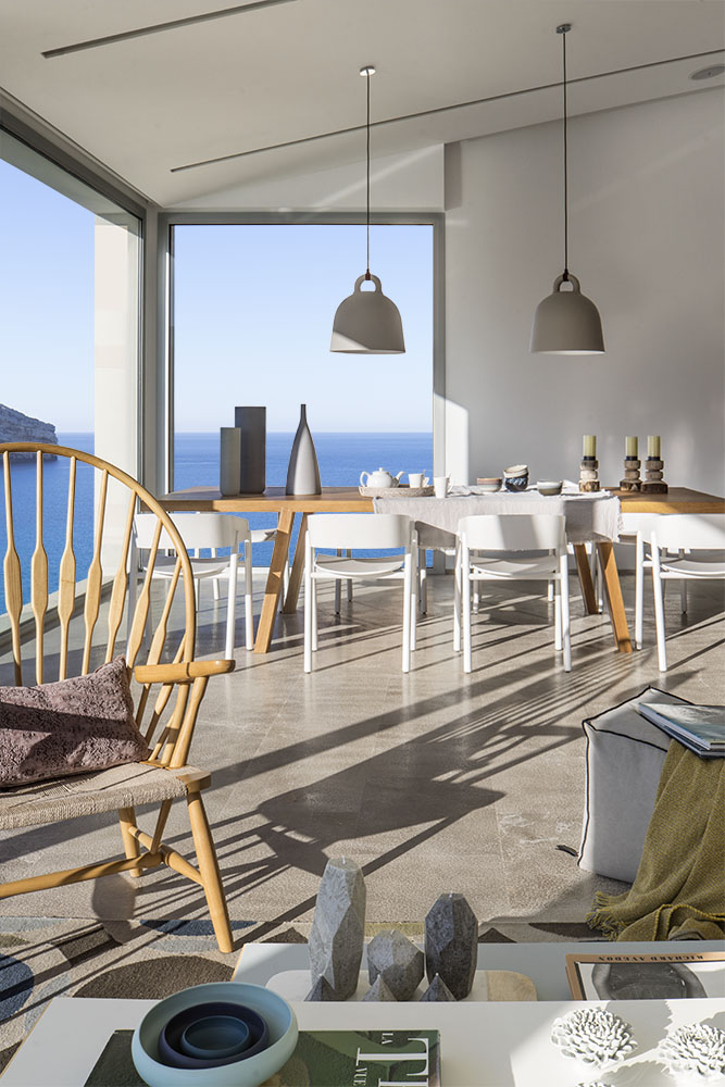 Vista desde el salón hacia el comedor con cristalera panorámica, mesa de madera y mar azul